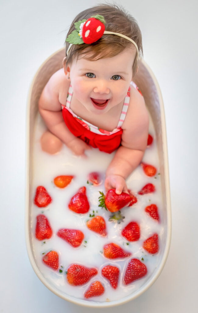 Baby one-year-old in a strawberry milk bath smiling up at the camera during a berry first birthday themed photoshoot